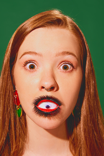 Shocked woman with eye makeup on lips against green background. Psychological thriller on self-perception and identity. Concept of pop art photography, creativity, surrealism, horror, quirky