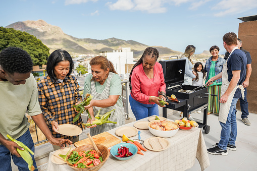 Multigenerational people doing barbecue at home's rooftop - Multiracial friends having fun eating and cooking together during weekend day - Summer and food concept - Main focus on asian woman face