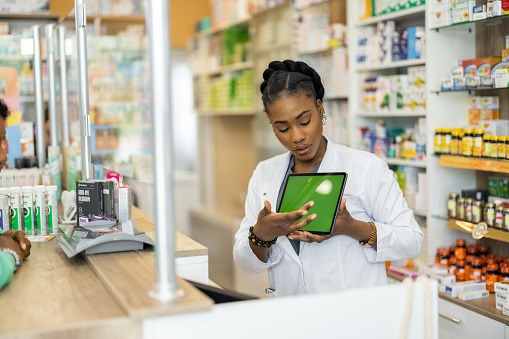 Young African female pharmacist standing behind the counter and showing products on the digital tablet to customer