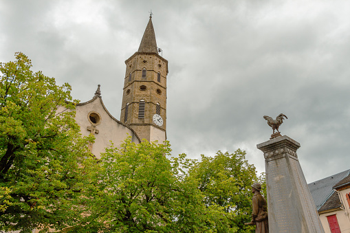 View of the tower of the church of Massat, Ariège, Occitanie, France. Village houses. Memorial to the dead soldiers of the first world war in the foreground, with the French rooster symbol.