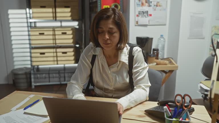 Mature hispanic woman detective working at computer in police department office, focused and serious.