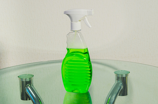 A green bottle of cleaner sits on a table. The bottle is clear and has a white cap. The table is made of glass and has a silver frame