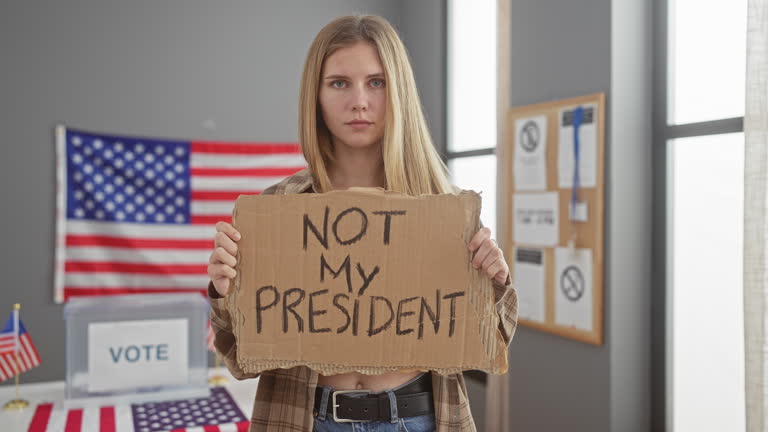A serious young blonde woman holds a protest sign in an american voting station.