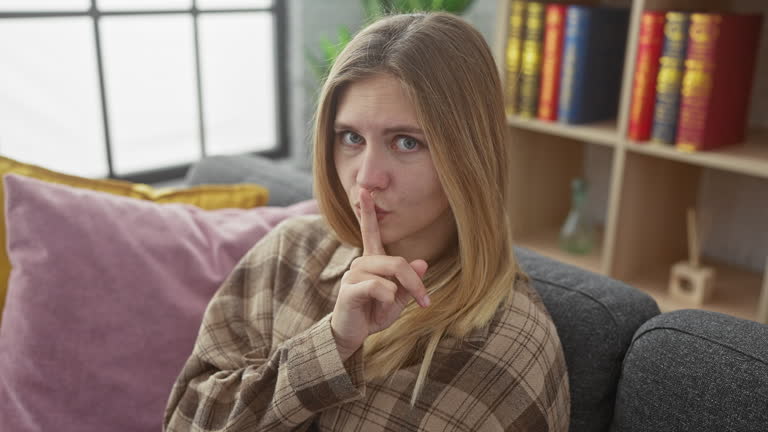 Hush! beautiful young blonde woman gesturing for silence, a secret sign with finger on lips, sitting on home sofa in calm room, quiet indoor portrait.