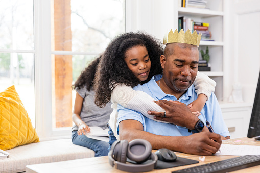 The mid adult father wears his paper crown and continues to work while daughter puts her arms around him.