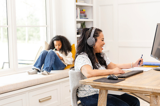 The focus of the photo is on a mid adult mother attending a virtual meeting as her elementary age daughter completes her homeschool assignment in the background.