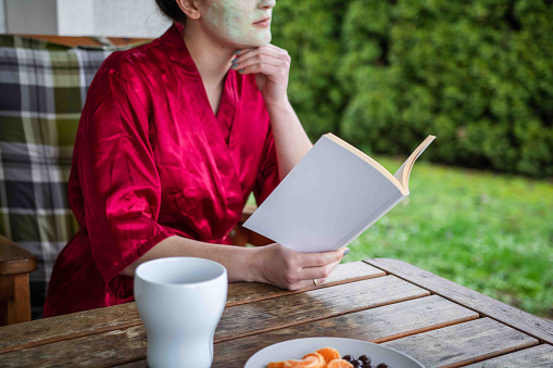 A woman wrapped in a red spa robe enjoys reading a blank book, embodying relaxation and self-care at home.