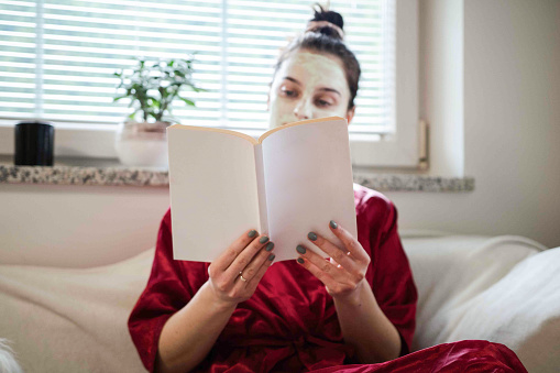 A woman wrapped in a red spa robe enjoys reading a blank book, embodying relaxation and self-care at home.