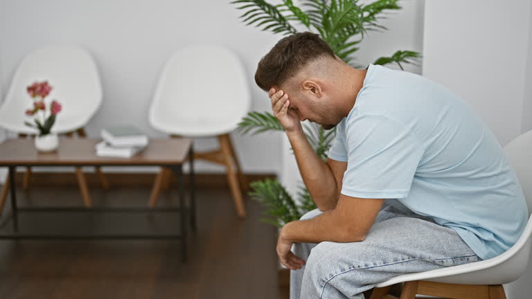 A distressed young hispanic man sitting in a clean modern indoor waiting room featuring white chairs and plants.