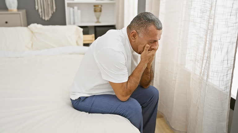 A distressed middle-aged hispanic man sitting on a bed in a well-lit bedroom, expressing worry or sadness.