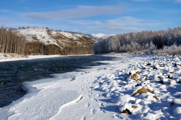 A river flows with ice along its shore, framed by a majestic mountain in the background. The sky is filled with clouds, creating a beautiful natural landscape. Russia, Chuisky ridge, Katun river.