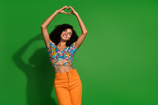 Photo of nice young woman curly hair make heart gesture looking up head cute surprise for anniversary isolated on green color background.