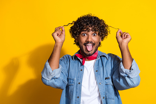 Photo of attractive young man hold curly strands tongue out dressed stylish denim clothes red scarf isolated on yellow color background.