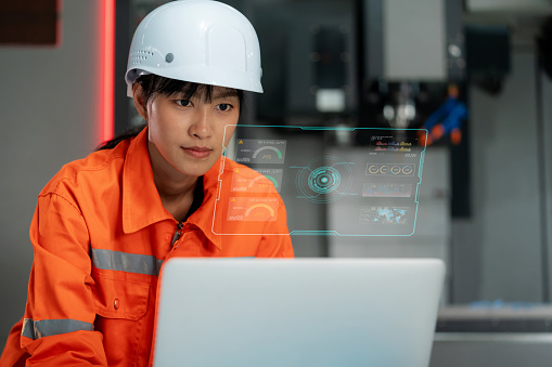 Young engineer is deeply focused on analyzing data using a laptop and viewing information on a graphical user interface in a modern factory setting. Dressed in a hardhat and reflective clothing, the engineer demonstrates expertise and precision in utilizing technology for efficient manufacturing processes.