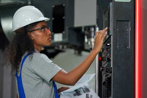 A skilled female engineer is operating a CNC machine in a modern factory. Wearing a hardhat and reflective clothing for safety while pressing buttons on the control panel with precision and expertise. Dedicated professional showcases efficiency, accuracy, and a commitment to sustainable energy and zero waste in the manufacturing industry.
