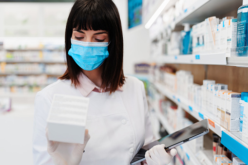Beautiful pharmacist with protective face mask working and standing in a drug store and doing a stock take. Portrait of a positive healthcare worker or a chemist at his work.