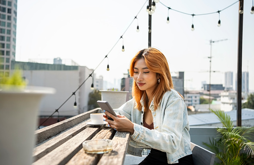 Beautiful young asian woman using her mobile phone while sitting at rooftop cafe table with coffee