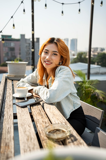 Portrait of pretty young asian woman sitting at table of rooftop cafe and looking at camera with smile on face