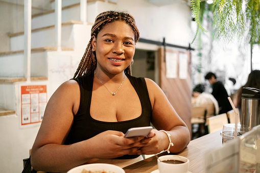 Portrait of beautiful young african woman using her mobile phone while sitting at cafe table with coffee