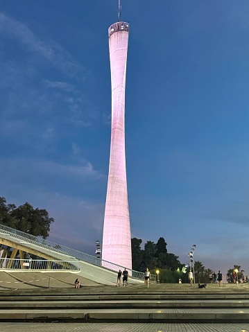 A tall white building with a pink top is lit up at night. The building is surrounded by trees and people are walking around it