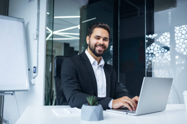 istock Modern Office Portrait of Stylish Hispanic Businessman Work on Laptop. makes Data Analysis, Looks at Camera and Smiles. Entrepreneur Works on e-Commerce Startup Project 2135684506