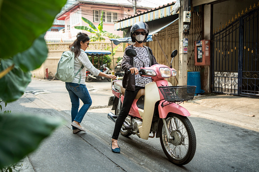 Senior woman wearing a protective face mask and a helmet waiting for a female passenger to get on the back of her motorbike for a ride in the city