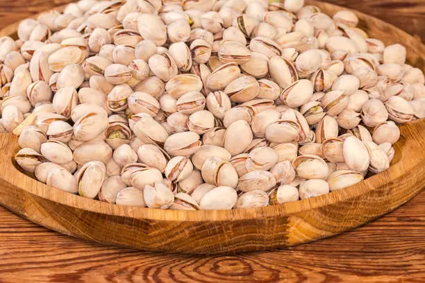 Heap of the roasted salted pistachio nuts with partly naturally open shells on a big wooden dish on the old rustic table, fragment close-up in selective focus