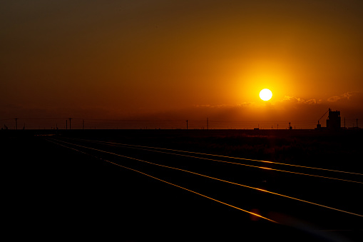 A breathtaking sunset dominates the horizon, casting an intense warm glow while silhouetting roadside structures and power lines. Parallel railway tracks glistening in the fading light guide the viewer's eye toward the distance.