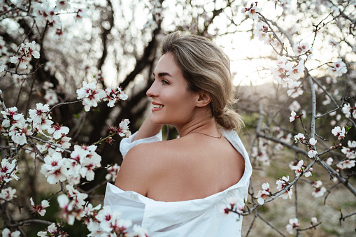 Beautiful young woman wearing a white shirt, elegantly posing near the blossoming almond trees.