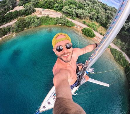 A mid adult man taking a selfie with wide angle camera, he climbed the sailboat mast and is enjoying the view from above, on Meganisi island, Greece
