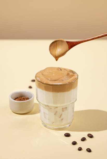 istock Dalgona coffee. Whipped instant coffee with ice and milk on a yellow background with shadow. Foam drips from the spoon into the glass. 2135912603