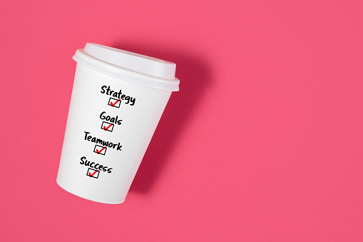 Conceptual shot with paper coffee cup and business related words; strategy, goals, teamwork, success as a choice on it on pink background