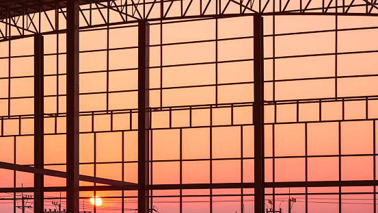 Silhouette metal industrial factory building structure in construction site against orange sunset sky background