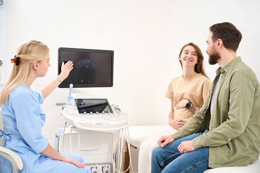 Female gynecologist showing screen with result of ultrasound scanning of woman patient pregnant belly to young smiling european couple looking at each other in clinic. Pregnancy and paternity