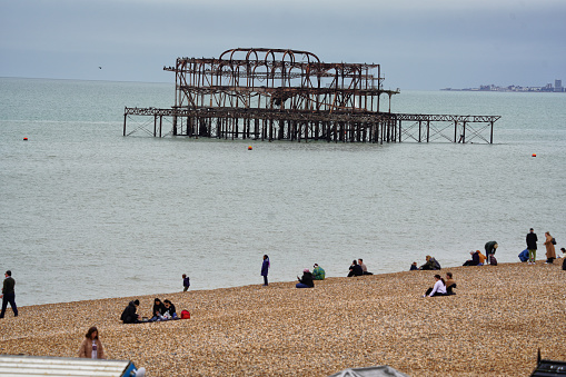 Brighton Beach of Brighton, Brighton And Hove, East Sussex, Southeastern England, United Kingdom, Britain, Europe - 16th March 2024: Brighton West Pier ruins; Atmospheric remnants of a once bustling 19th-century pleasure pier, destroyed by fire in 2003. Located at Kings Rd, Brighton BN1 2FL, United Kingdom. Beach, Coast, Seaside & Shore.

On a cloudy spring day, crowds stroll along the iconic Kings Road, a seaside street in the heart of Brighton and Hove, this major location buzzes with cultural hubs at the seafront. There, locals and visitors alike bask on the pebbled beach, soaking in the stunning coastal scenery of the English Channel meeting this quintessential British seaside town. 

Some sit enjoying the beach & nature and ocean air, while others explore the fishing village ambience near the water's edge, capturing the perfect blend of urban and natural beauty in East Sussex. 

A non-urban landscape, prime viewpoints, relaxing outdoors along the coast, Brighton Beach, seaside culture and scenic splendour in England, Europe.