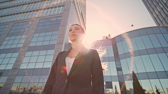 Posh businesswoman standing sunlight looking at distance bottom view. Serious beautiful woman boss posing in front modern town skyscrapers. Confident lady in elegant suit outdoor. Camera moving around