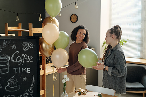 Happy young African American woman with bunch of balloons looking at colleague with dreadlocks while both decorating cafe after refit