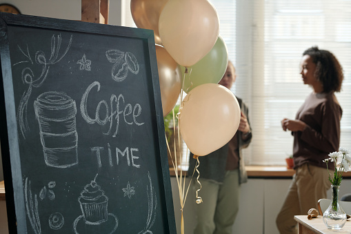 Bunch of balloons decorating chalkboard with advert drawing of cup of coffee and cupcake standing against two young female employees