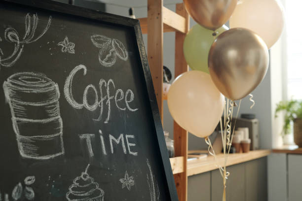 Blackboard with advert or offer presented by cup of coffee drawn with white chalk against bunch of beige and golden balloons and counter