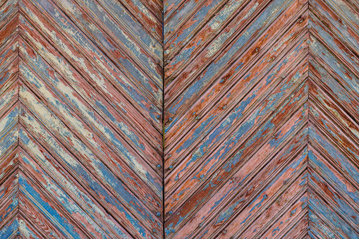 Artistic old wooden planks gate chevron pattern with peeled brown and blue paint layers under sun-bleached blue paint layer. Full-frame flat background and texture.