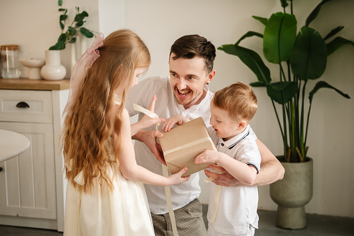 Kids make surprise for Fathers Day. Daughter and son greet father and open present gifts at home. The child congratulates surprised daddy closeup. Children embrace smiling dad. Happy family holidays.