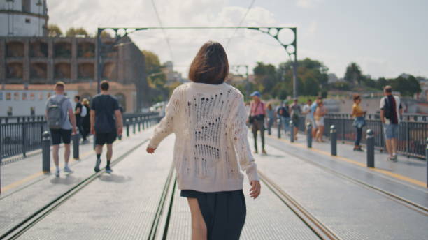 Stylish woman walking rails at summer city back view. Relaxed brunette girl going overcrowded street in beautiful cityscape. Calm model spinning alone looking camera at tram ways. City life concept