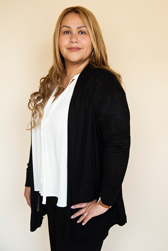 Portrait of stylish modern professional hispanic woman on beige background. She is in her forties, wearing a white blouse and a black cardigan over a black pants, standing and looking at the camera. Vertical waist up studio shot with copy space. This was taken in Montreal, Quebec, Canada.