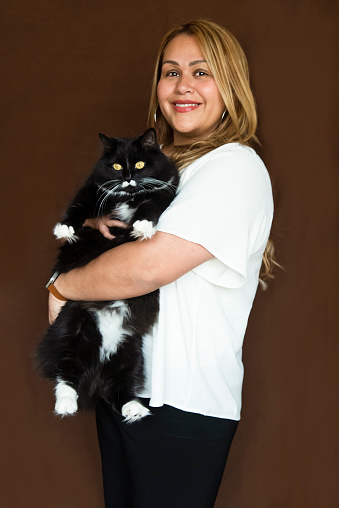 Portrait of stylish modern professional hispanic woman holding a tuxedo cat on brown background. She is in her forties, wearing a white blouse over a black pants, standing and looking at the camera. Vertical waist up studio shot with copy space. This was taken in Montreal, Quebec, Canada.