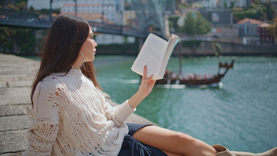 Romantic girl reading novel at river bank zoom out. Focused woman enjoying weekend hobby resting in warm sunlight. Beautiful lady holding book literature study outdoors. Relaxation leisure concept