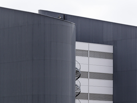 Abstract daytime close-up of the exterior of a modern industrial building with facade cladding in light and dark surfaces and a spiral metal staircase in the middle
