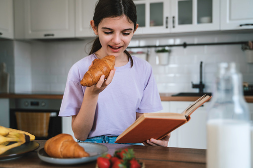Beautiful girl reading a book and having breakfast in the kitchen.