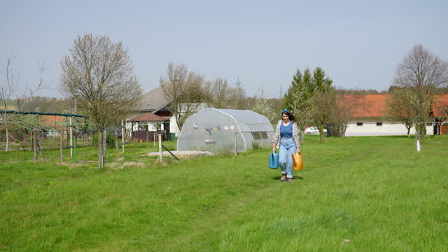 SLO MO Happy Young Woman Gardener Walking with Watering Cans on Grassy Field on Sunny Day