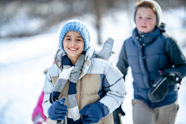 A small group of school-aged children are seen running in the snow together on a Winter's day.  They are each dressed warmly in layers, coats, and mittens as they play together on the cool day.