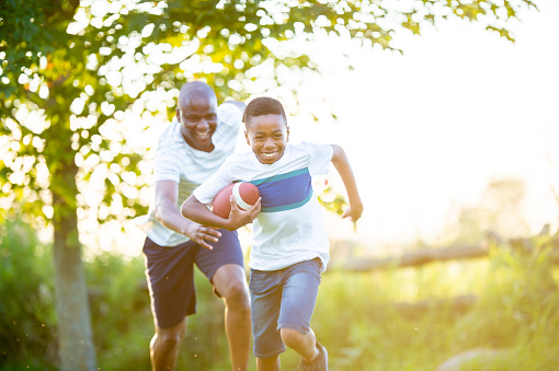 A young boy of African decent runs with a football tucked under his arm as he plays a quick game with his Father during a summer family BBQ.  The two are dressed casually and smiling as they enjoy the time spent together.